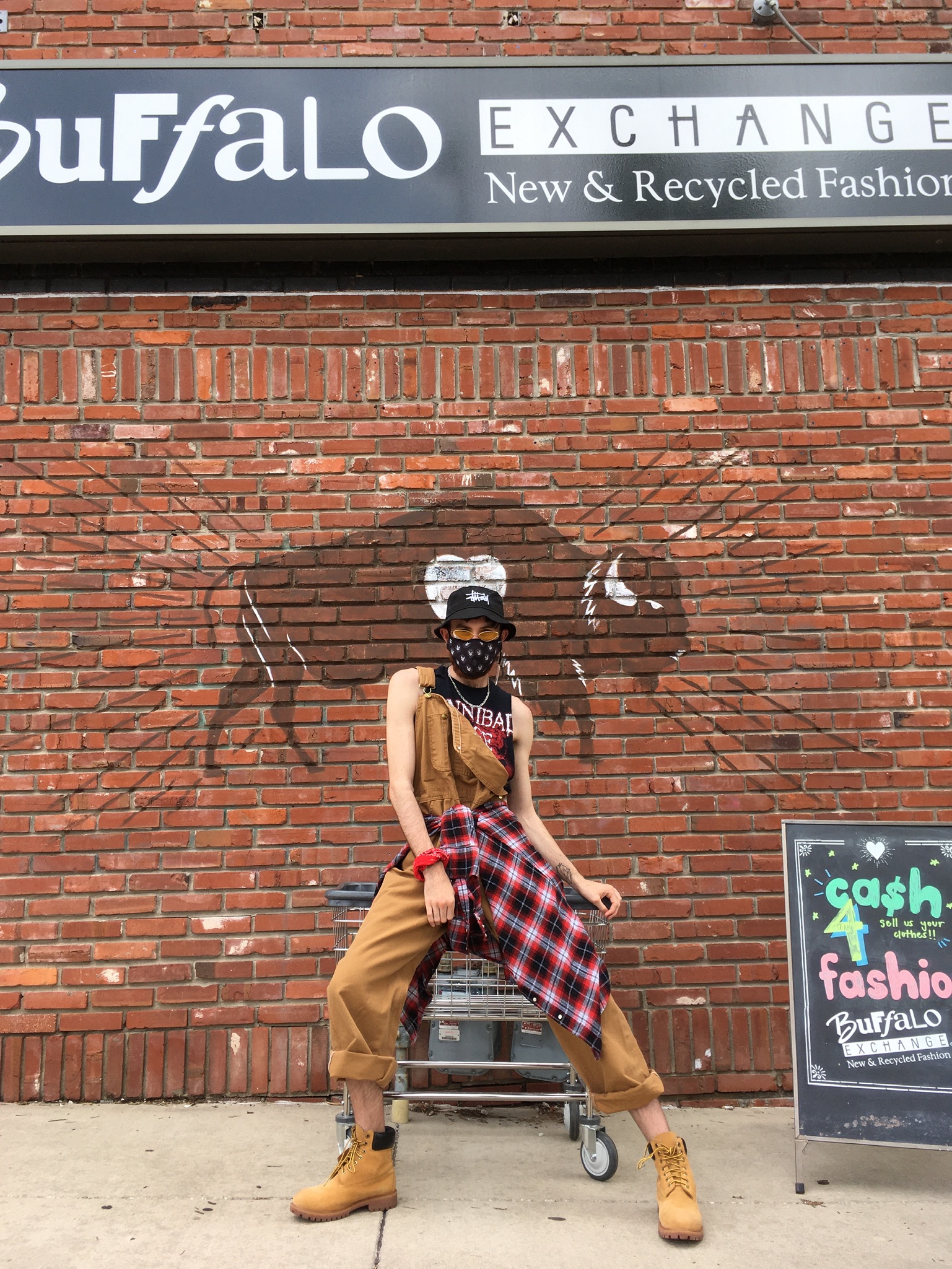 Person in front of a buffalo mural wearing Carhartt tan overalls, a plaid button down tied around their waist, and Timberland boots