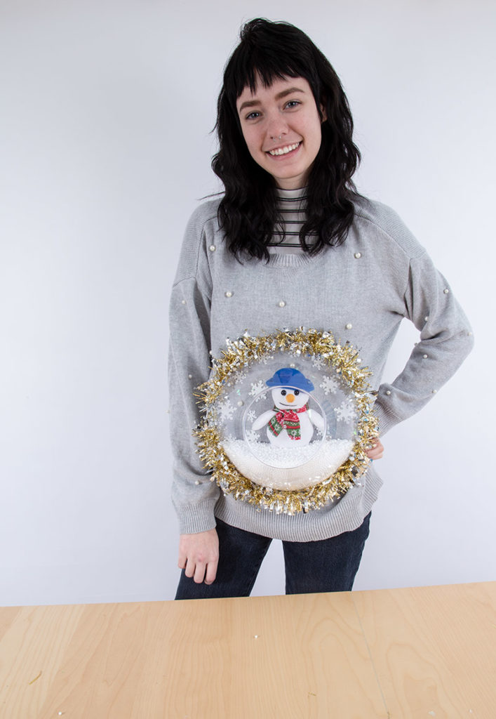 Person wearing a DIY holiday sweater with a plastic bowl and snowman stuffed animal glued to the front making it look like a snowglobe