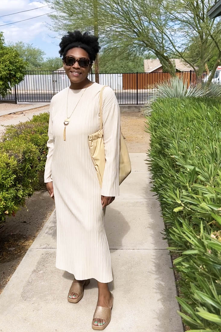 Person standing on sidewalk wearing floor-length beige dress and gold sandals