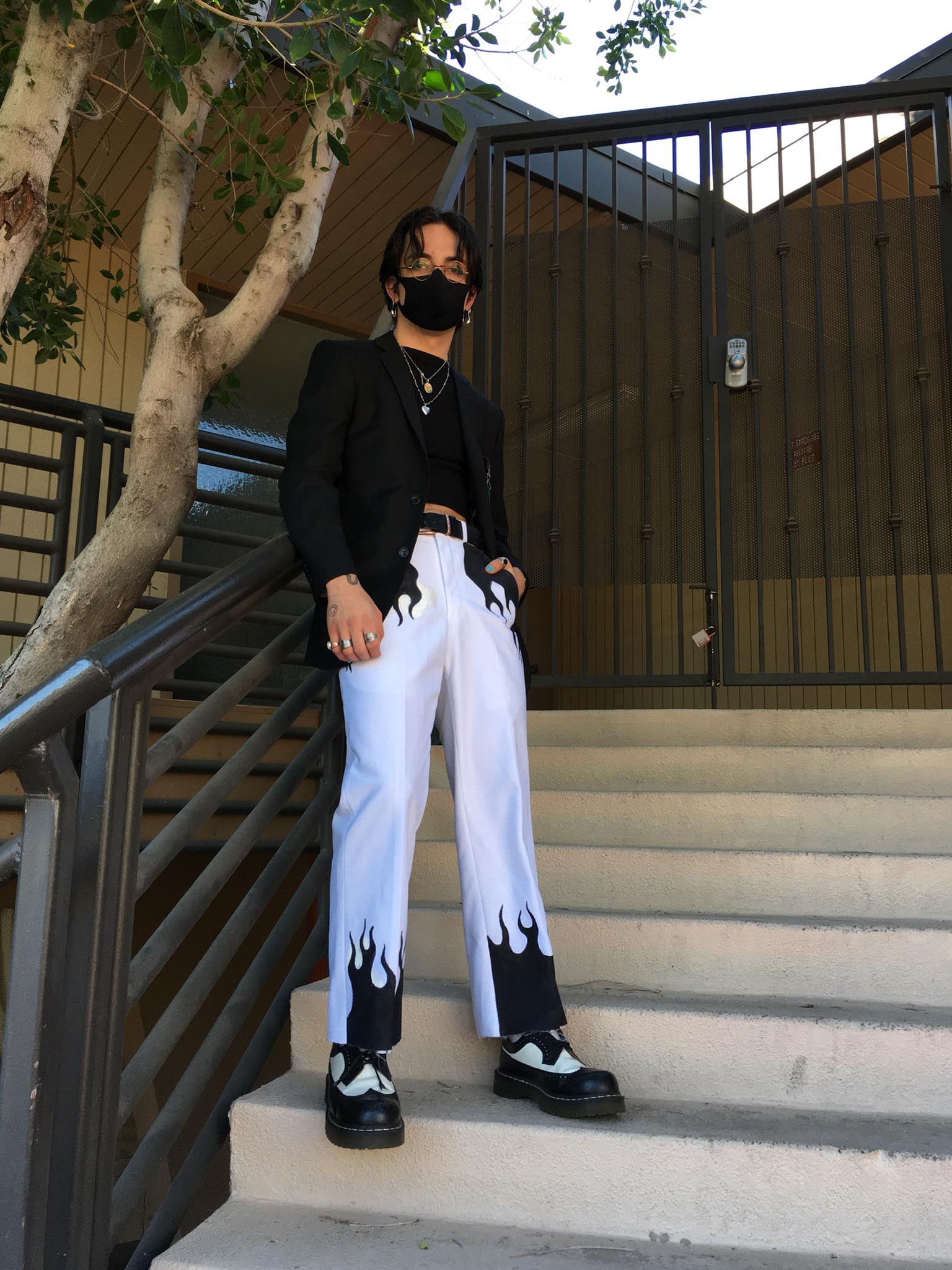 Person standing on steps with tree in background wearing white pants with black flame details and black top and jacket