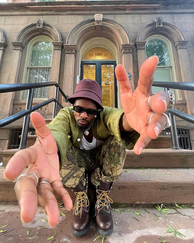 Customer @_boobmiles wearing a brown bucket hat, olive green quilted jacket, camo pants, silver jewelry, and brown lace up boots.