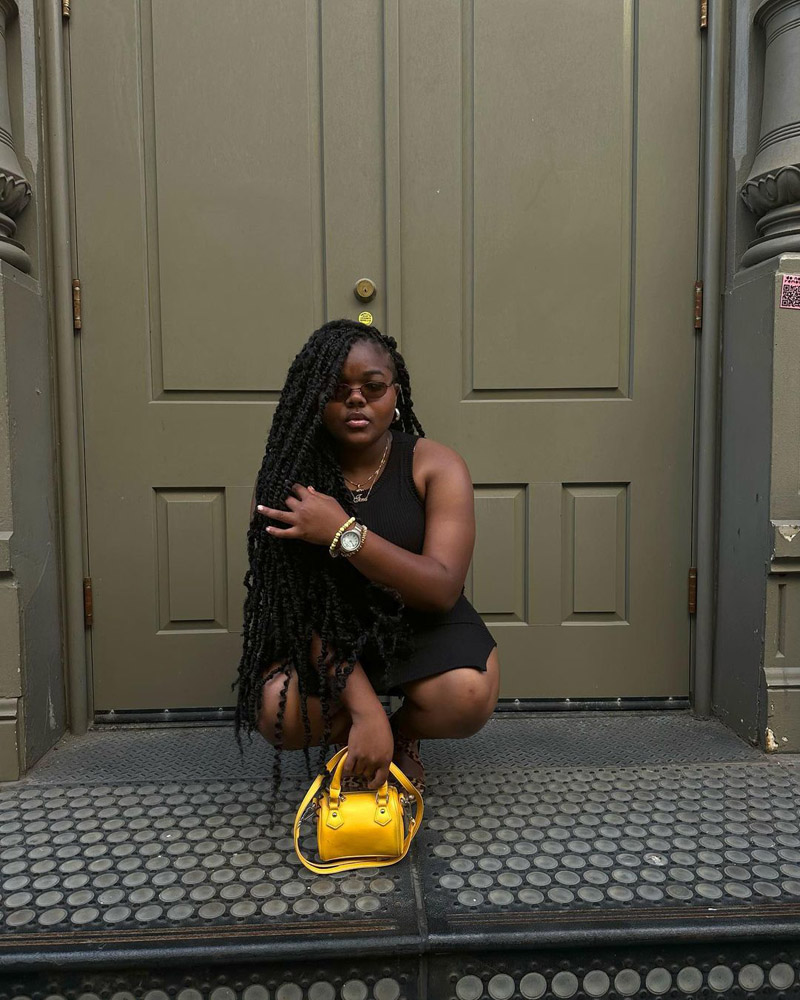 @tinaashanti wearing a black mini dress, gold necklaces, a gold watch, and a bright yellow mini purse