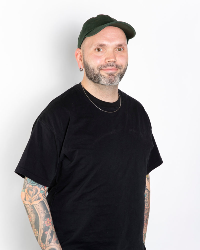 A headshot of Mike wearing a green baseball cap, black tee shirt, and a chain necklace