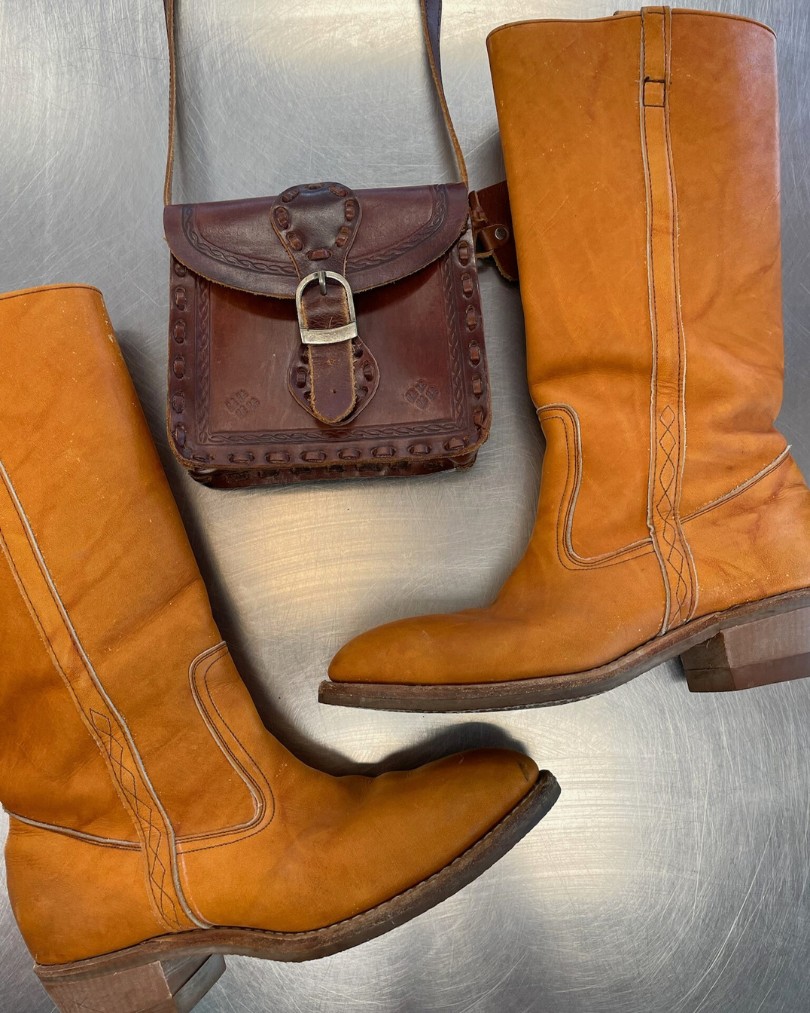 Flat lay of vintage western boots in tan leather with a mini tooled leather crossbody purse at the Buffalo Exchange buy counter Flat lay of vintage western boots in tan leather with a mini tooled leather crossbody purse at the Buffalo Exchange buy counter