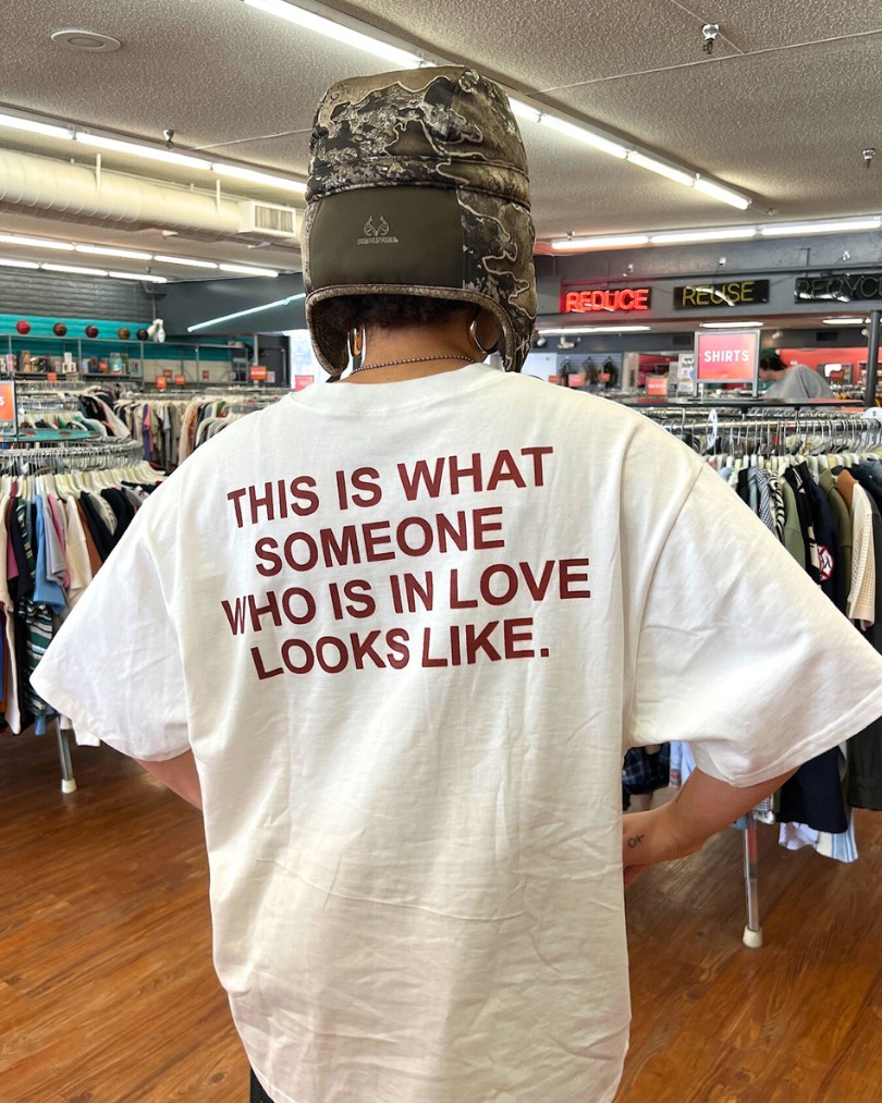 A person inside Buffalo Exchange showing the back of their graphic tee shirt that says in capitalized red letters: "This is what someone who is in love looks like." A person inside Buffalo Exchange showing the back of their graphic tee shirt that says in capitalized red letters: "This is what someone who is in love looks like."
