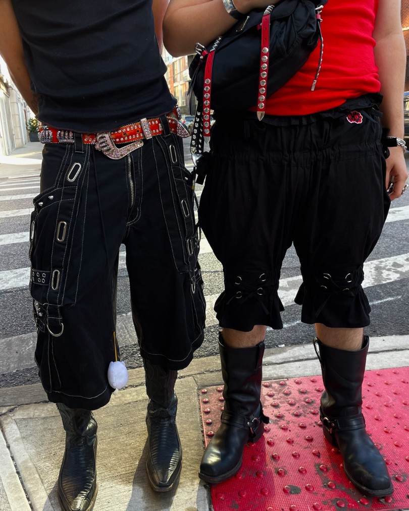 A close up of two people dressed in red & black moto style on a New York City street corner wearing tanks, shorts with grommet details, and leather harness boots A close up of two people dressed in red & black moto style on a New York City street corner wearing tanks, shorts with grommet details, and leather harness boots