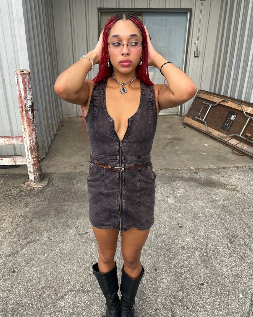 Person standing in front of an industrial building while wearing metal framed glasses, an acid wash denim dress, leather knee-high moto boots and silver jewelry Person standing in front of an industrial building while wearing metal framed glasses, an acid wash denim dress, leather knee-high moto boots and silver jewelry