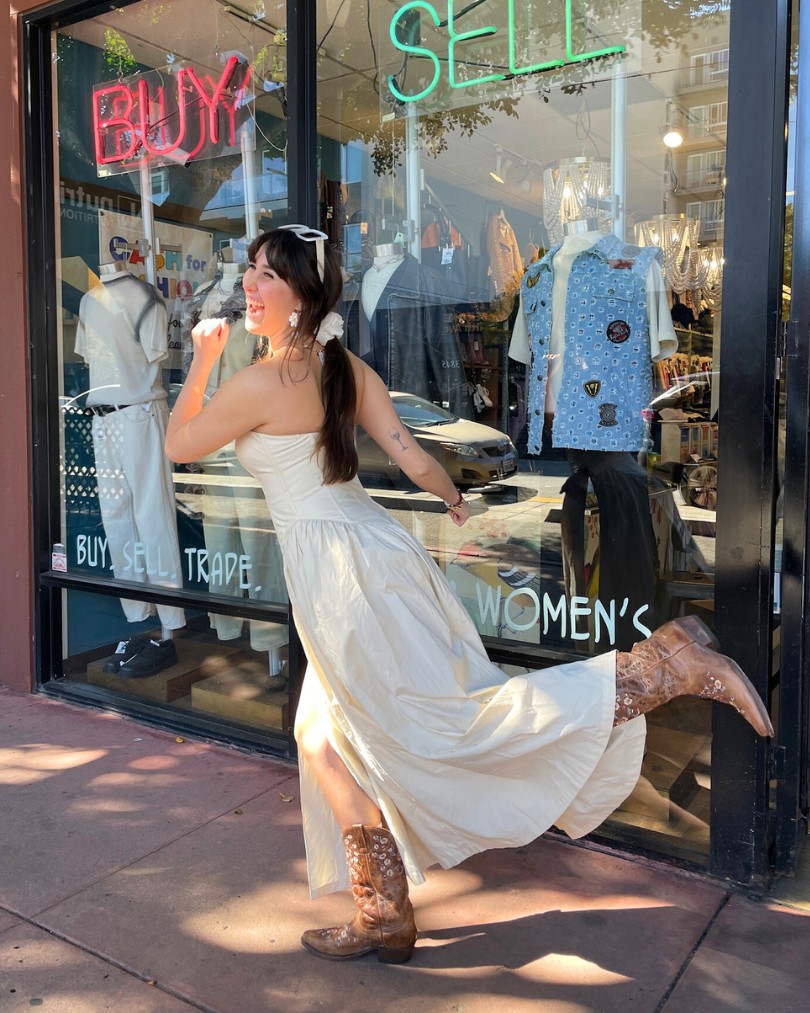Person in front of neon signs that say "Buy" and "Sell" pretending to run into a Buffalo Exchange while wearing a strapless cream dress and brown leather western boots Person in front of neon signs that say "Buy" and "Sell" pretending to run into a Buffalo Exchange while wearing a strapless cream dress and brown leather western boots